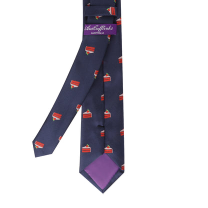 The Strawberry Cake Skinny Tie is a navy blue tie with a repeating pattern of red cake slices topped with white frosting and cherry, plus a purple inner lining—perfect for fans of fun food-themed ties.