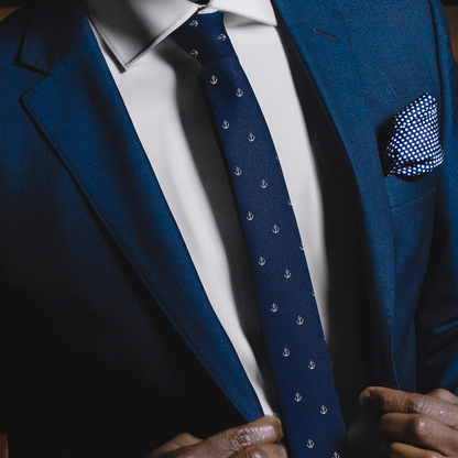 Close-up of a person wearing a dark blue suit with a white shirt. They are adjusting an Anchor Skinny Tie, embodying stability, and have a blue and white patterned pocket square.