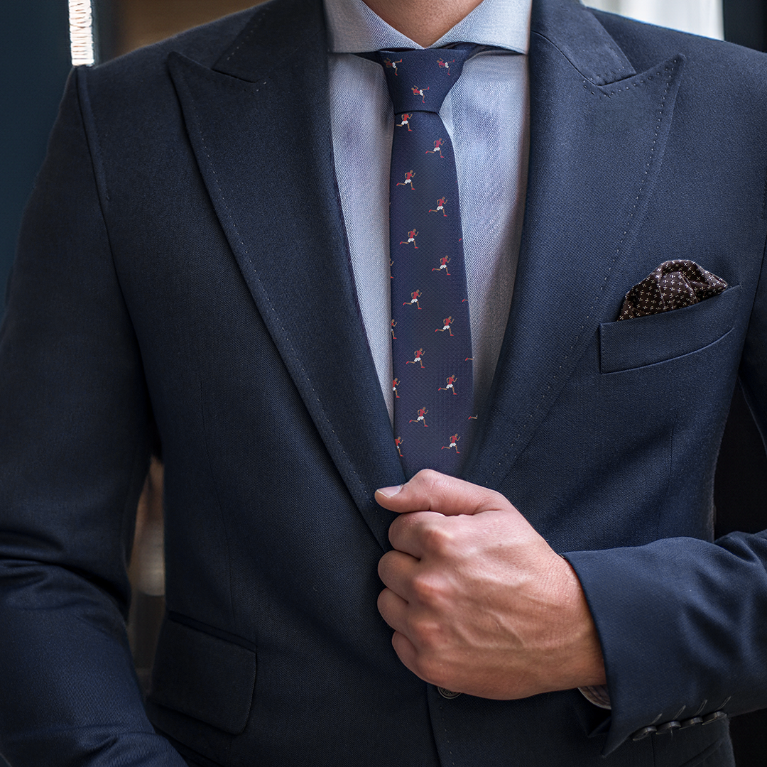 A person in a navy blue suit holds their lapel with one hand, wearing a blue dress shirt and an Athletics Skinny Tie with red patterns. A polka-dotted pocket square peeks out from the suit's breast pocket, adding an extra touch of style.