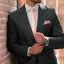 A person in a dark suit and pink tie, exuding pastel sophistication, adjusts their cuff while leaning against a brick wall and showcasing a watch and the Baby Pink Skinny Necktie and Pocket Square Set.