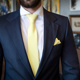 A person in a navy blue suit with a white shirt, and a Baby Yellow Business Cotton Tie that pops alongside the matching pocket square, stands indoors.