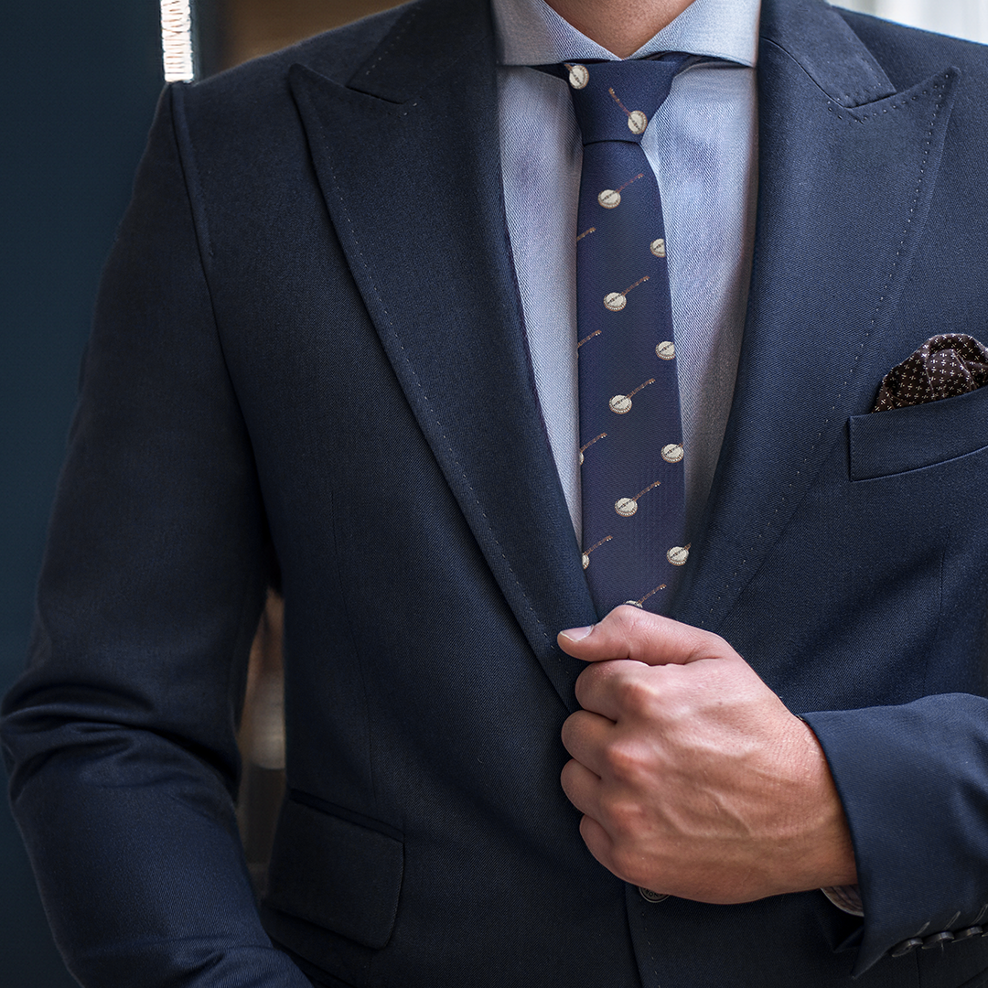 A person wearing a dark blue suit with a Banjo Skinny Tie and a polka-dot pocket square adjusts their jacket, exuding an air of sophistication.