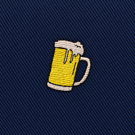 Embroidered patch of a frothy beer mug on dark blue diagonal fabric background, perfect for accenting the Beer Skinny Tie.