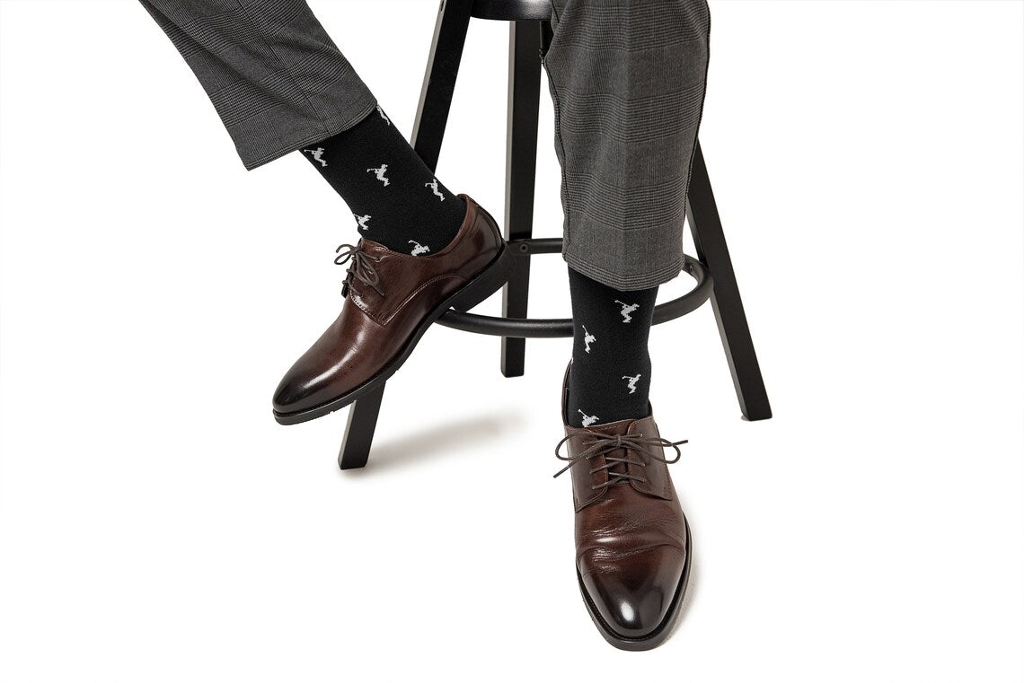 A person wearing dark brown dress shoes, Golf Swing Black Socks, and gray plaid pants sits on a black stool against a white background.