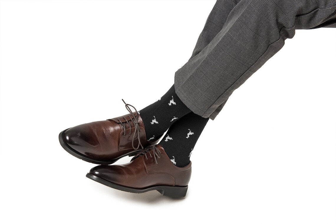 A person wearing gray dress pants, Golf Swing Black Socks with white dog patterns, and brown leather dress shoes sits cross-legged against a white background.
