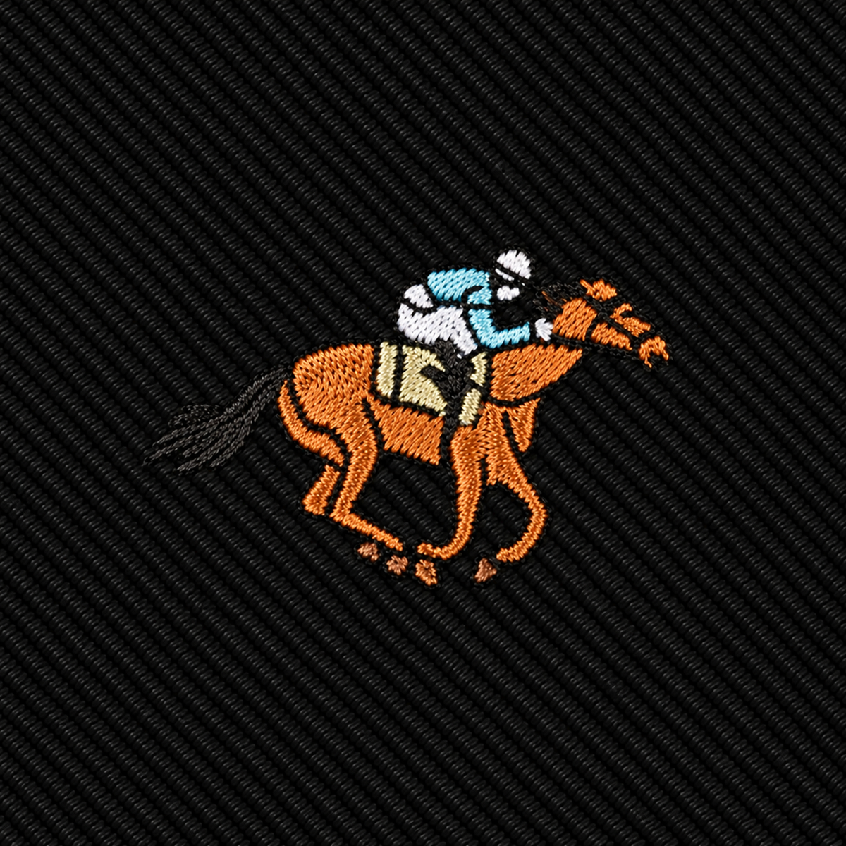 Embroidered image of a jockey in blue and white silks riding an orange horse, stitched on a dark, diagonal-patterned fabric background—perfect as a standout accent on the Horse Racing Skinny BLACK Tie or a unique addition to any horse racing tie collection.