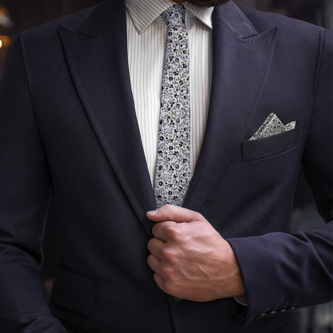 A person in a dark suit jacket, Black Print Floral Cotton Skinny Tie & Pocket Square Set with a sleek design adjusts their lapel.