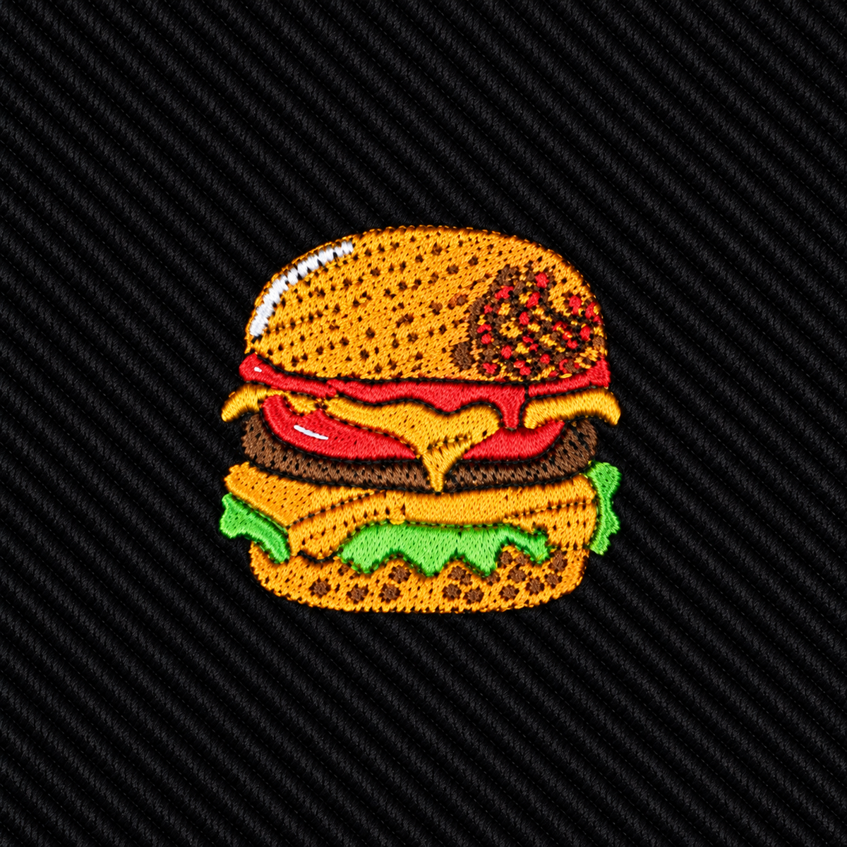 Introducing the Black Tie Burger: an embroidered patch featuring a cheeseburger with lettuce, tomato, cheese, and a sesame seed bun on a dark, textured background—an ideal gift for food lovers and the perfect quirky accessory for men’s fashion enthusiasts.