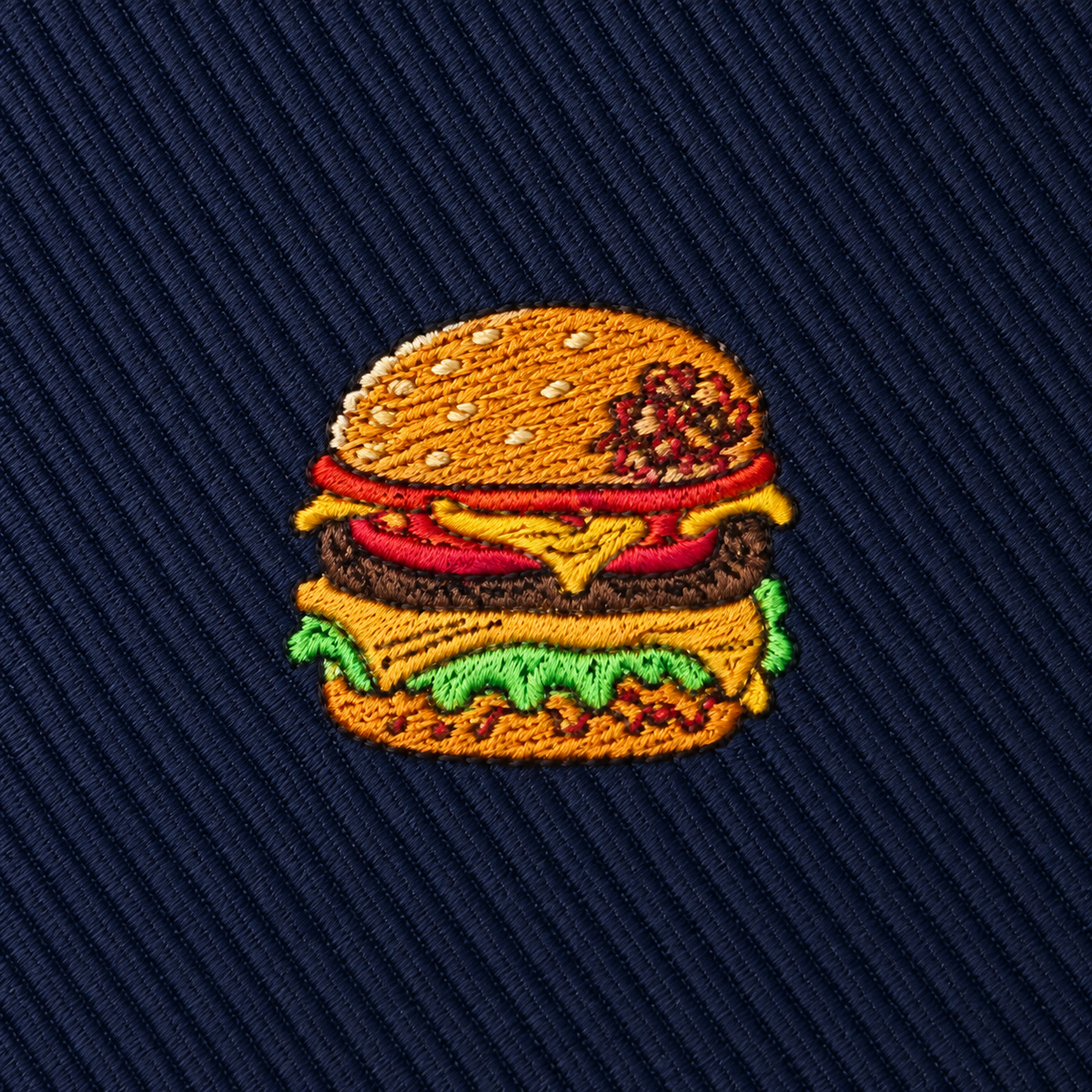Add a playful touch to your wardrobe with the Burger Skinny Tie, featuring an embroidered patch of a cheeseburger with lettuce, tomato, cheese, and a sesame seed bun on dark blue textured fabric—a perfect foodie gift or quirky accent for any burger lover.
