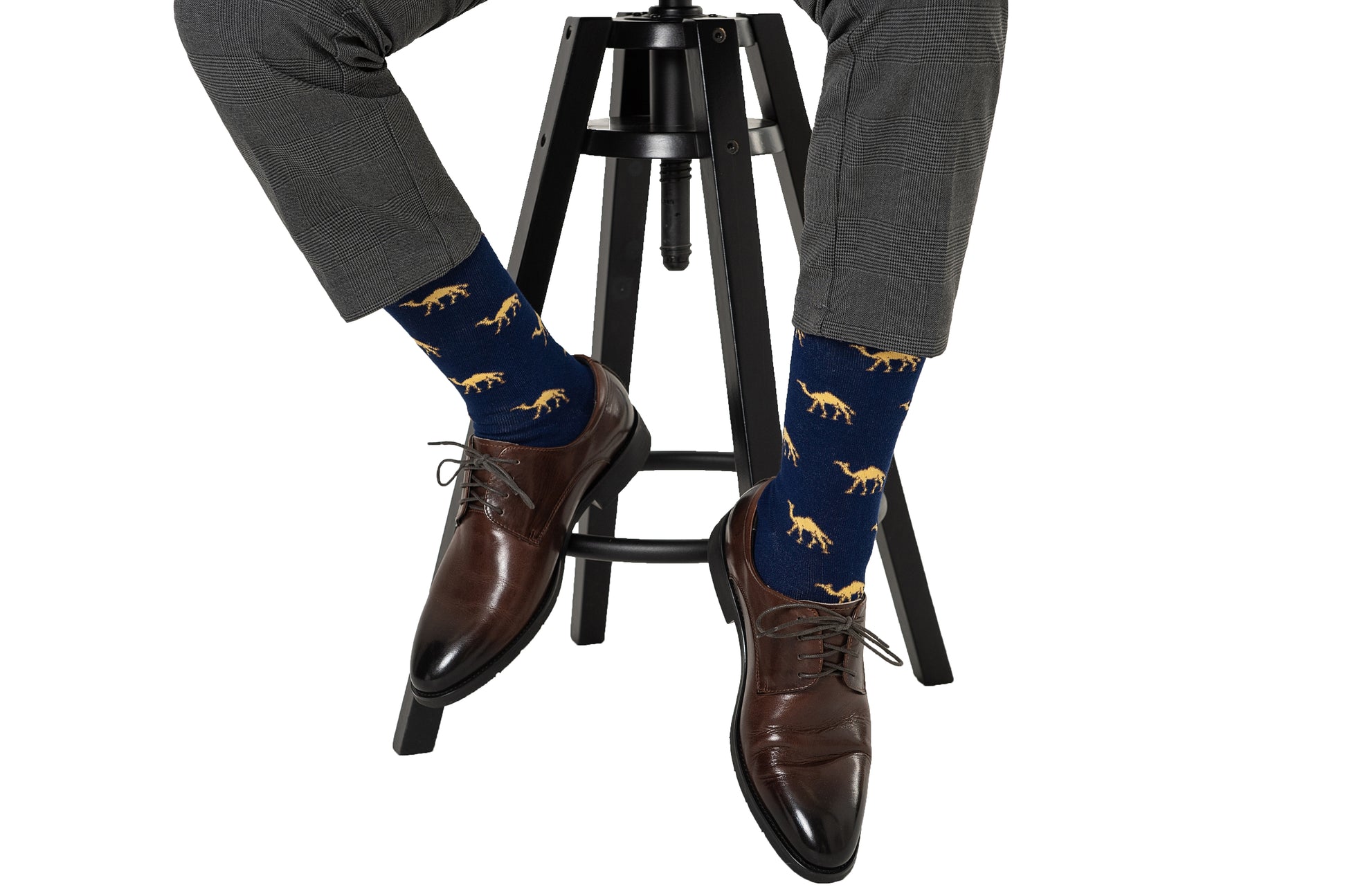 A person wearing gray plaid pants, brown dress shoes, and AusCufflinks Camel Socks—an ideal animal lover gift—seated on a black stool.
