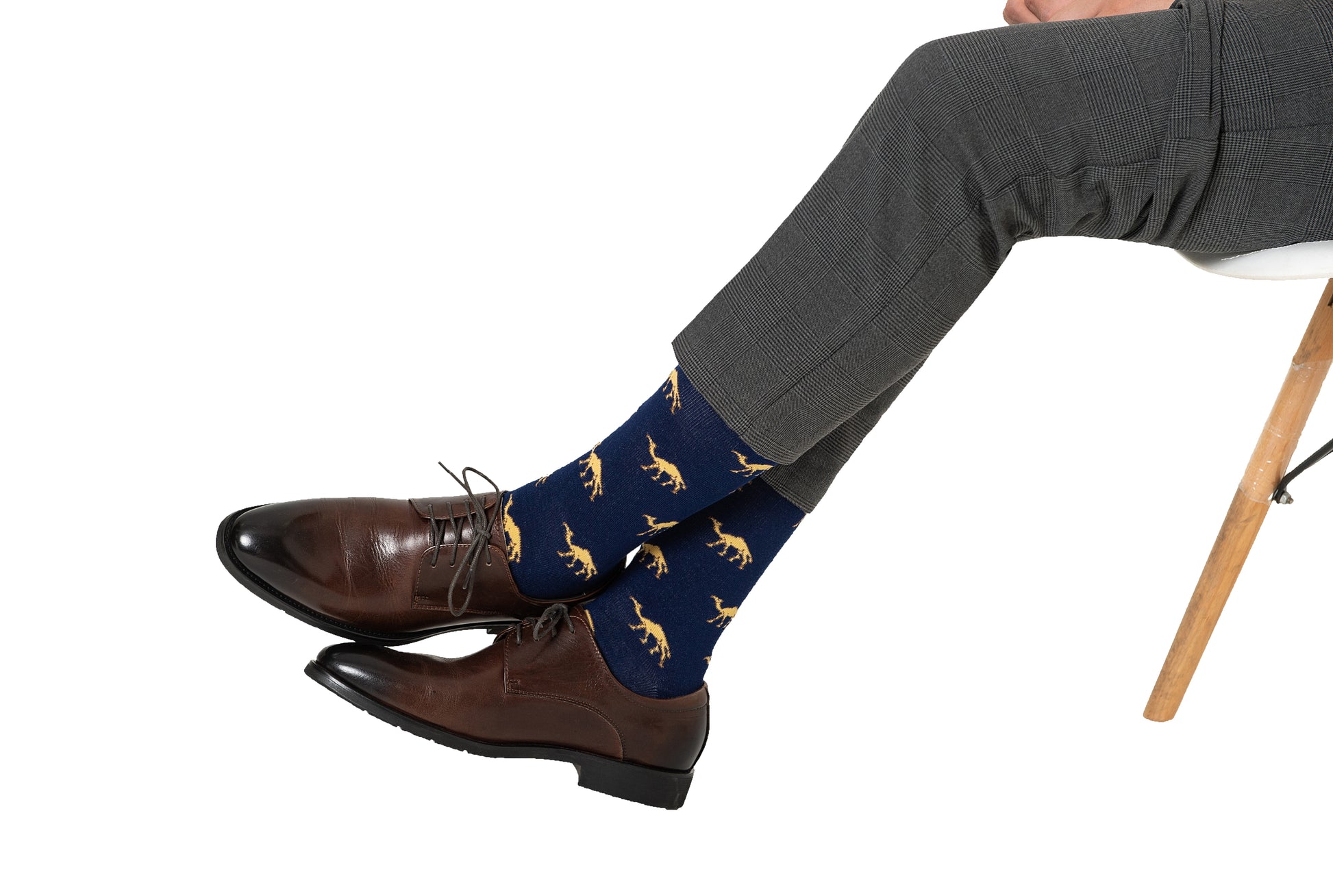 A person wearing gray dress pants, brown shoes, and Camel Socks with yellow dinosaur patterns sits cross-legged on a chair—a playful gift for animal lovers.