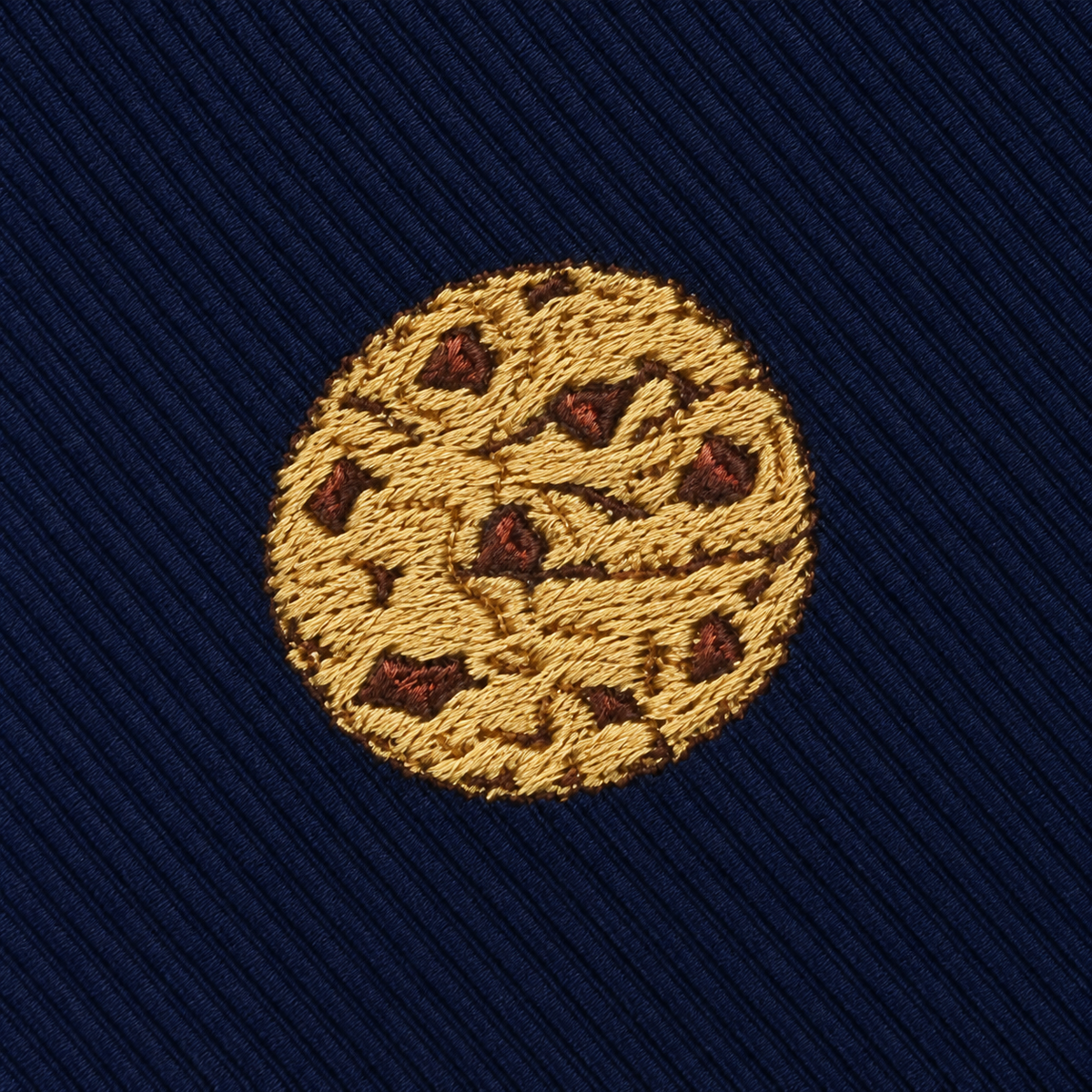 The Cookies Skinny Tie features a round, embroidered chocolate chip cookie motif on dark blue, diagonally ribbed fabric for a sweet, dessert-inspired look.