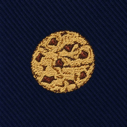 The Cookies Skinny Tie features a round, embroidered chocolate chip cookie motif on dark blue, diagonally ribbed fabric for a sweet, dessert-inspired look.