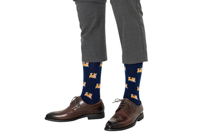 A person in gray plaid pants and brown shoes wears Corgi Dog Socks—navy socks with orange and white corgi designs—on a white background. Perfect for dog lovers or as a fun Corgi Dog Socks gift.