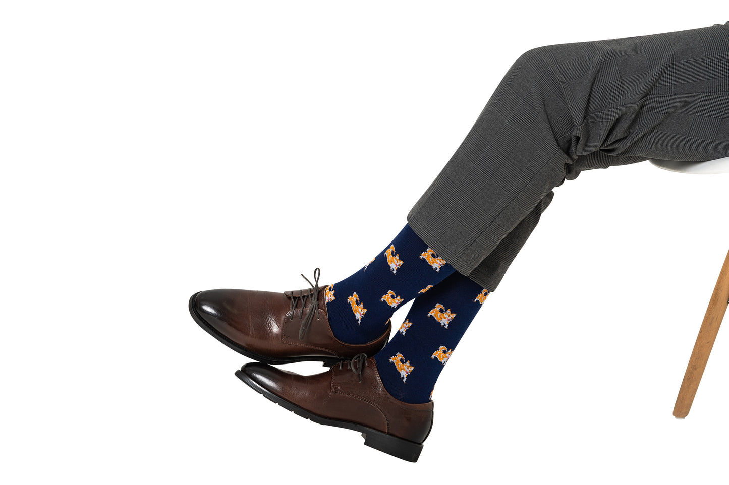 A person in gray dress pants, brown dress shoes, and Corgi Dog Socks featuring a playful dog design sits cross-legged on a white chair—an ideal style for any dog lover.