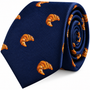 The Croissant Skinny Tie in navy blue features an embroidered pattern of small orange croissants and comes rolled up neatly—a gourmet-inspired fashion accessory that brings playful charm to any outfit.