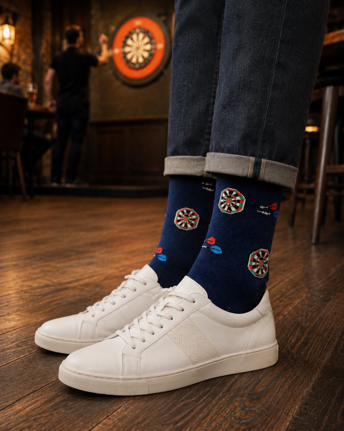 A person wearing white sneakers and Darts Socks featuring blue dartboard patterns stands on wooden floors with a dartboard visible in the background.