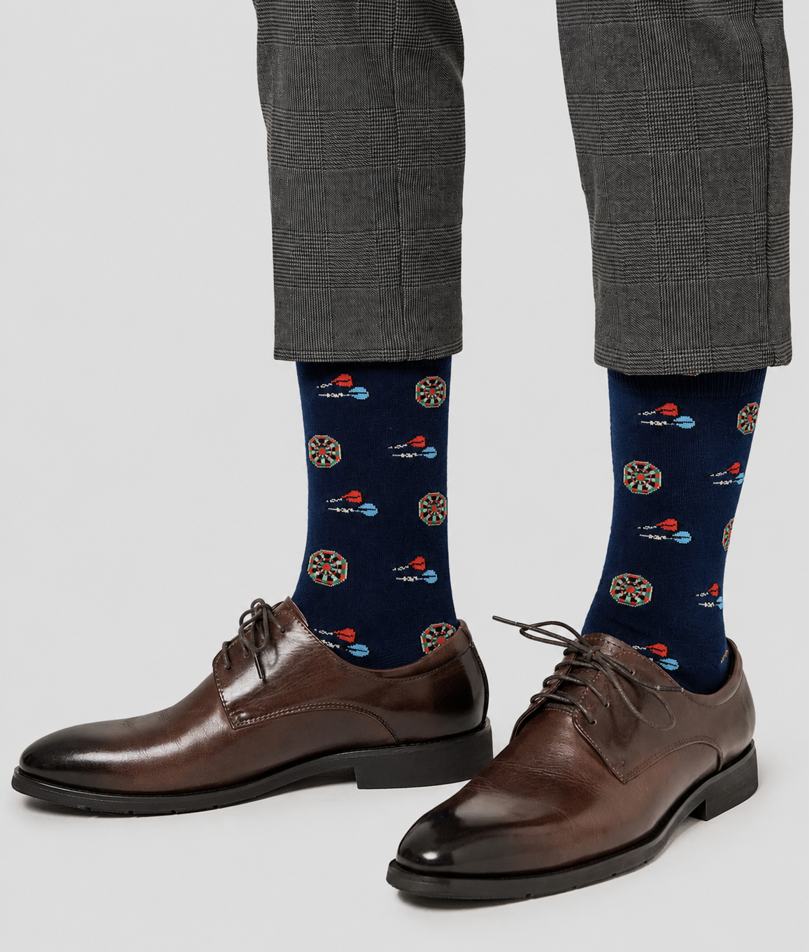 A person wearing dark brown dress shoes, gray plaid pants, and Darts Socks featuring dartboard and dart designs.