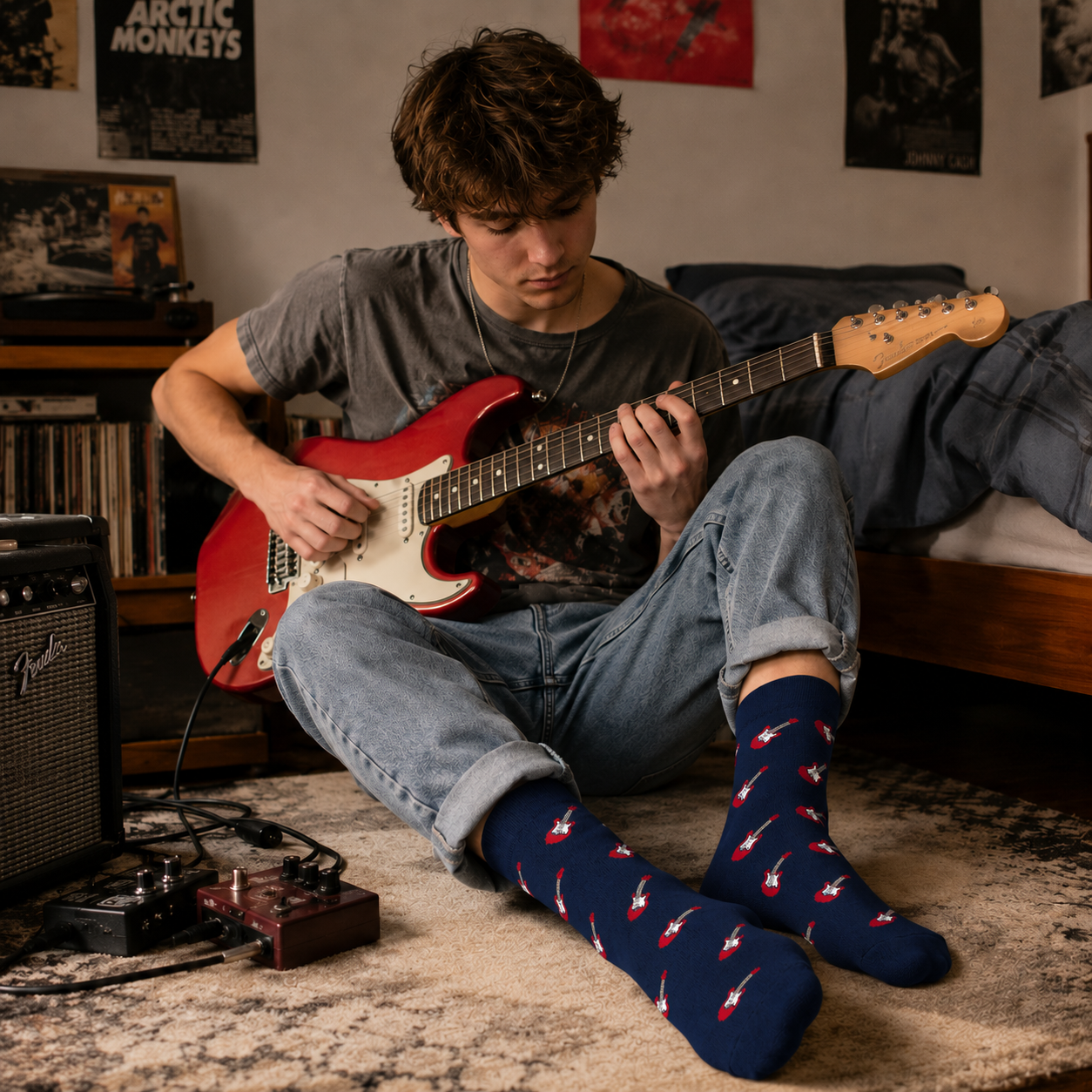 A young person sits on a bedroom floor playing a red electric guitar, wearing Electric Guitar Socks with blue fabric and guitar patterns, surrounded by music posters and equipment in the background.