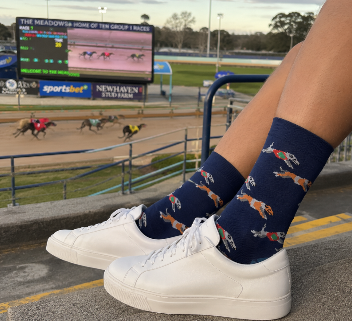 A person wearing white sneakers and Greyhound Racing Socks sits on steps at a racetrack, with a dog race and the scoreboard visible in the background.