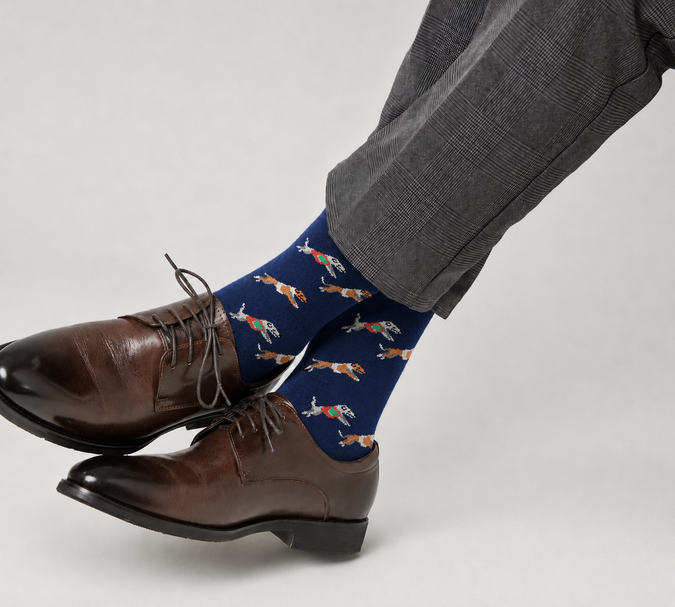 A person wearing brown dress shoes, gray plaid pants, and Greyhound Racing Socks with an orange fish pattern sits cross-legged.