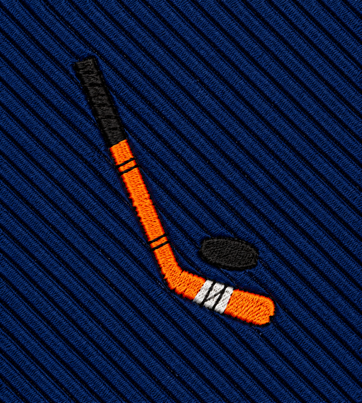 The Ice Hockey Skinny Tie features an embroidered orange hockey stick and black puck on blue, ribbed fabric, making it a standout accessory for ice hockey fans.