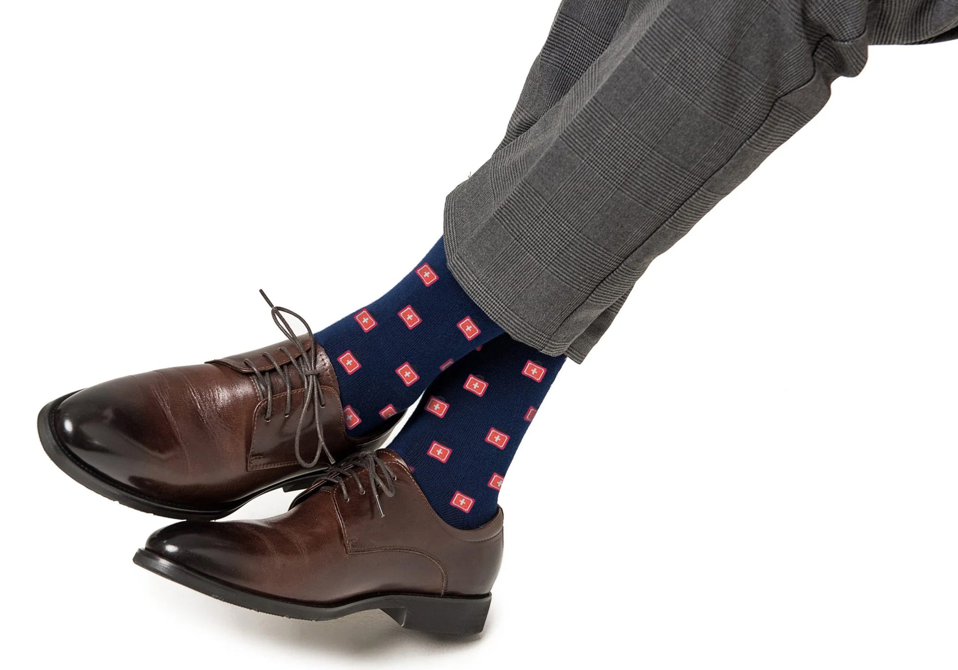 A person wearing brown leather dress shoes, Medical Cross Socks with a red cross pattern, and gray plaid dress pants is shown with legs crossed on a white background.