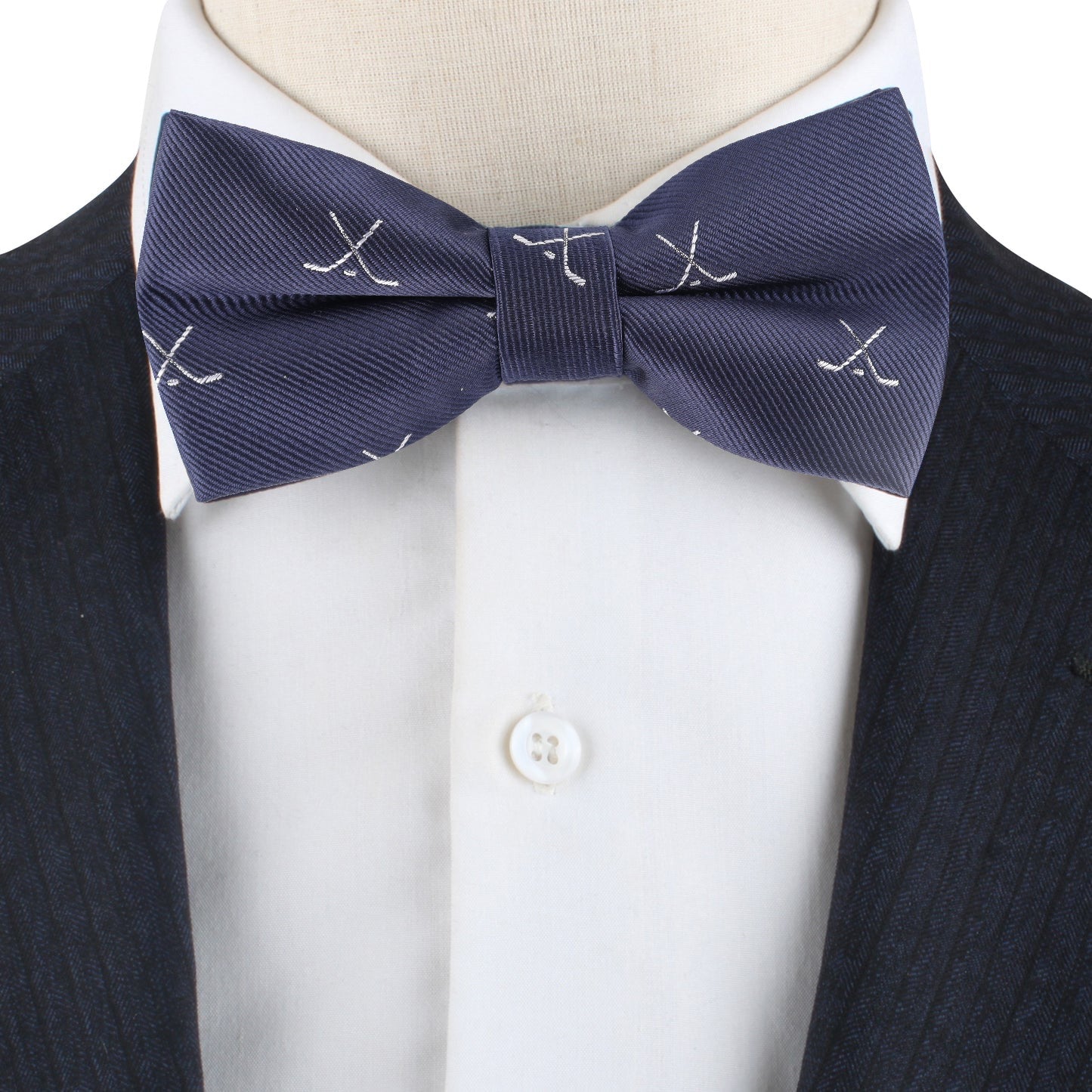 A close-up of a person wearing the Ice Hockey Cross Bow Tie with blue fabric, white embroidered crossed golf clubs, a white dress shirt, and a dark pinstriped suit jacket for a sporty touch.