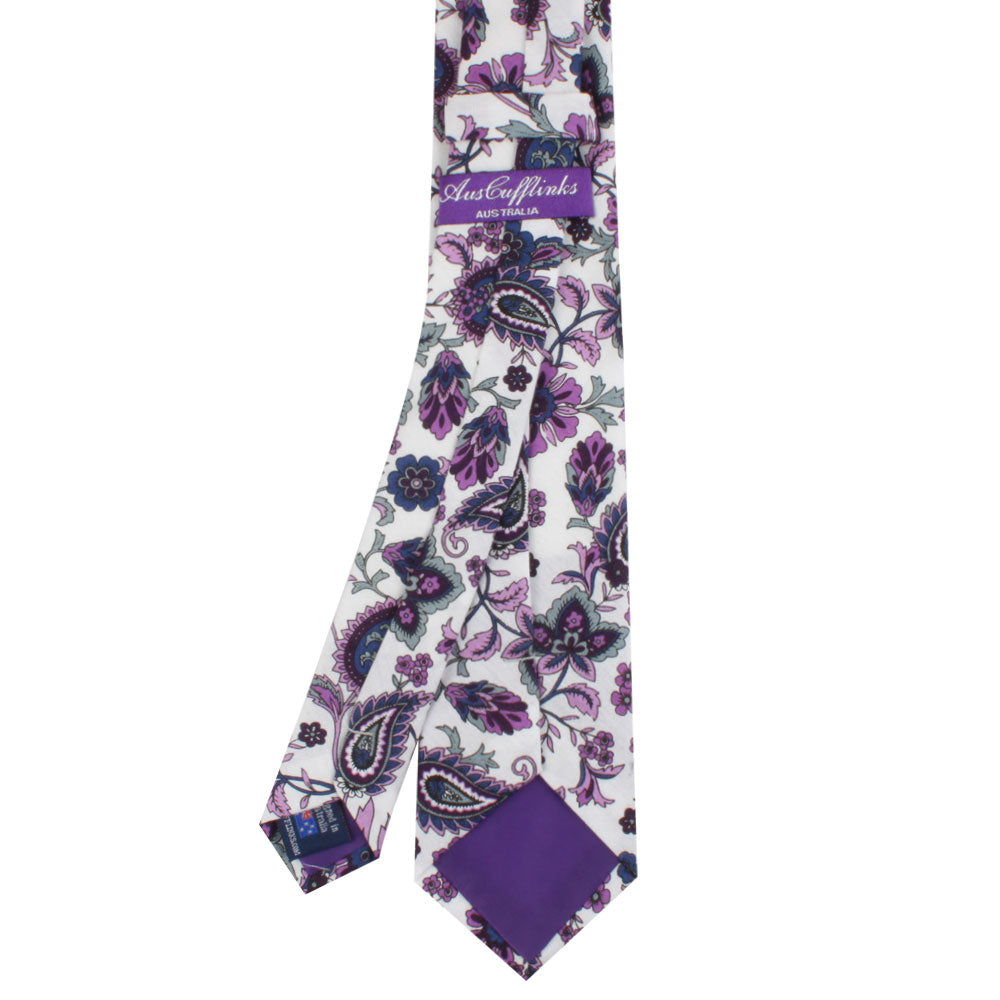 The Purple Floral White Paisley Skinny Tie features a slim design with purple and black floral paisley patterns, ideal for men’s formalwear or weddings; shown laid flat with its brand label on the back.