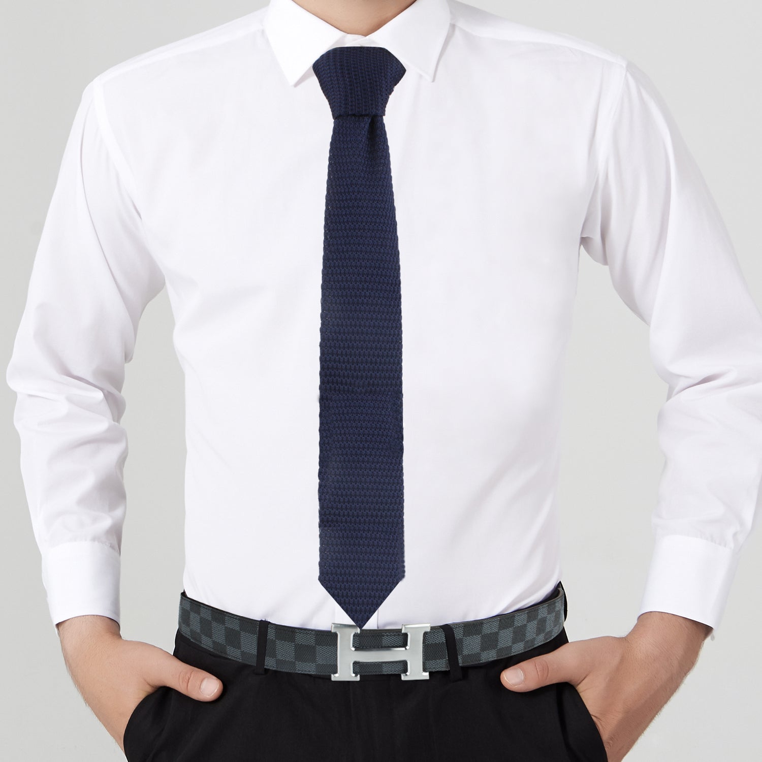 A person in a white dress shirt and Navy Knitted Skinny Tie, with hands in their pockets, exudes elegance, wearing a black checkered belt with an "H" shaped buckle.