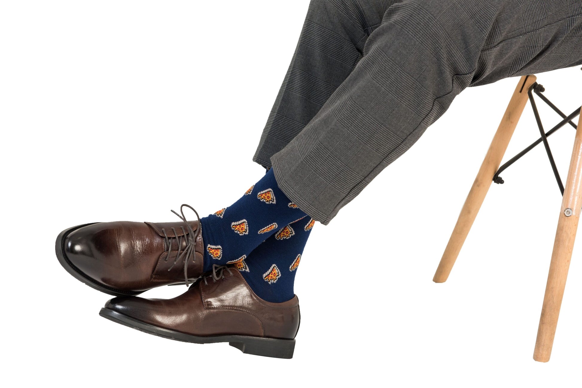 A person embodies comfort and style, wearing gray dress pants, brown leather shoes, and eye-catching Pizza Socks featuring vibrant orange and red pizza patterns while sitting with one leg crossed over the other on a wooden chair.