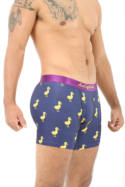 A man wearing Duck Underwear—navy blue boxer briefs adorned with a cheerful yellow duck pattern and featuring a vibrant pink waistband—stands against a white background, exuding plush comfort.