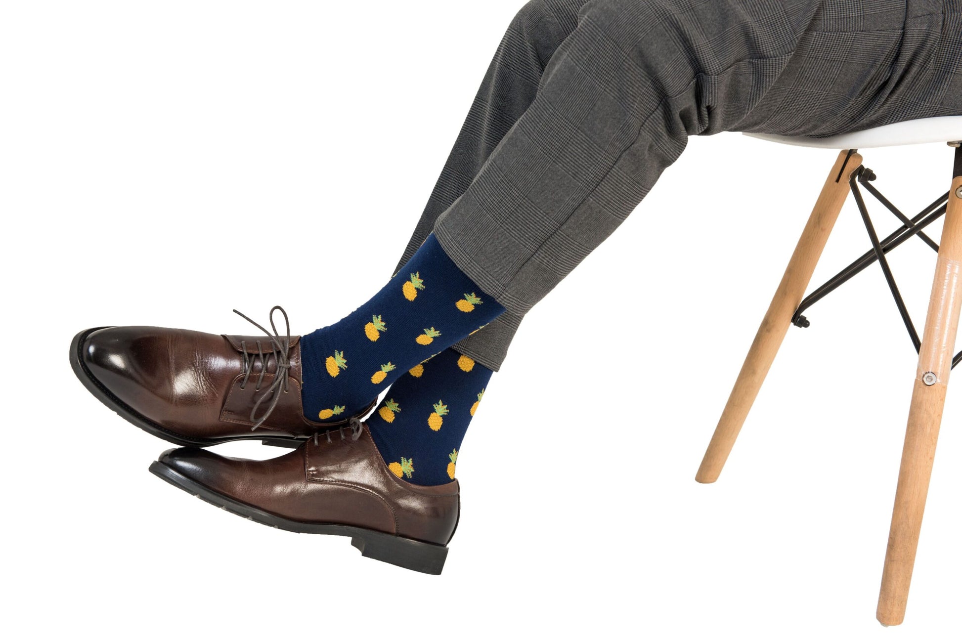 A person wearing gray pants and brown dress shoes sits on a white chair, showcasing Pineapple Socks. From heel to toe, the socks radiate tropical comfort.