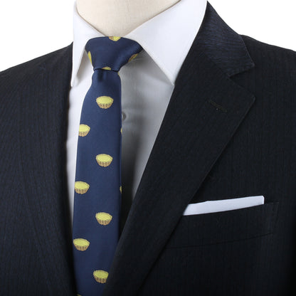 A close-up of a person in a dark suit wearing an Egg Tart Skinny Tie, featuring delightful yellow cupcake patterns, paired with a white dress shirt and complemented by a neatly folded white pocket square.