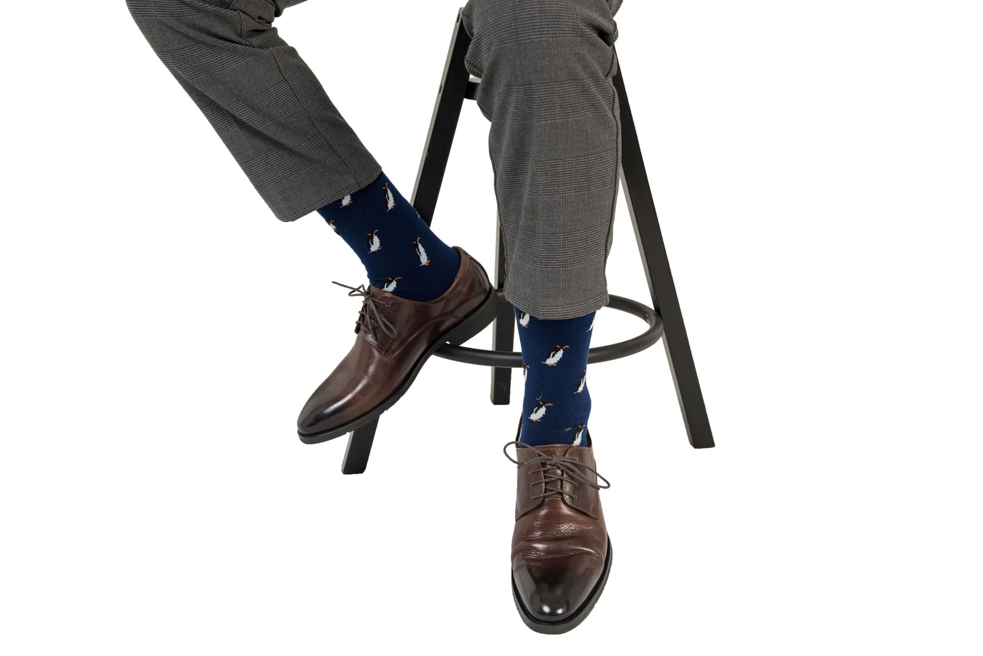 Man sitting on a stool, wearing grey trousers, brown dress shoes, and Penguin Socks—exuding an arctic allure while keeping it chill and comfortable. Only the lower half of the body is visible.