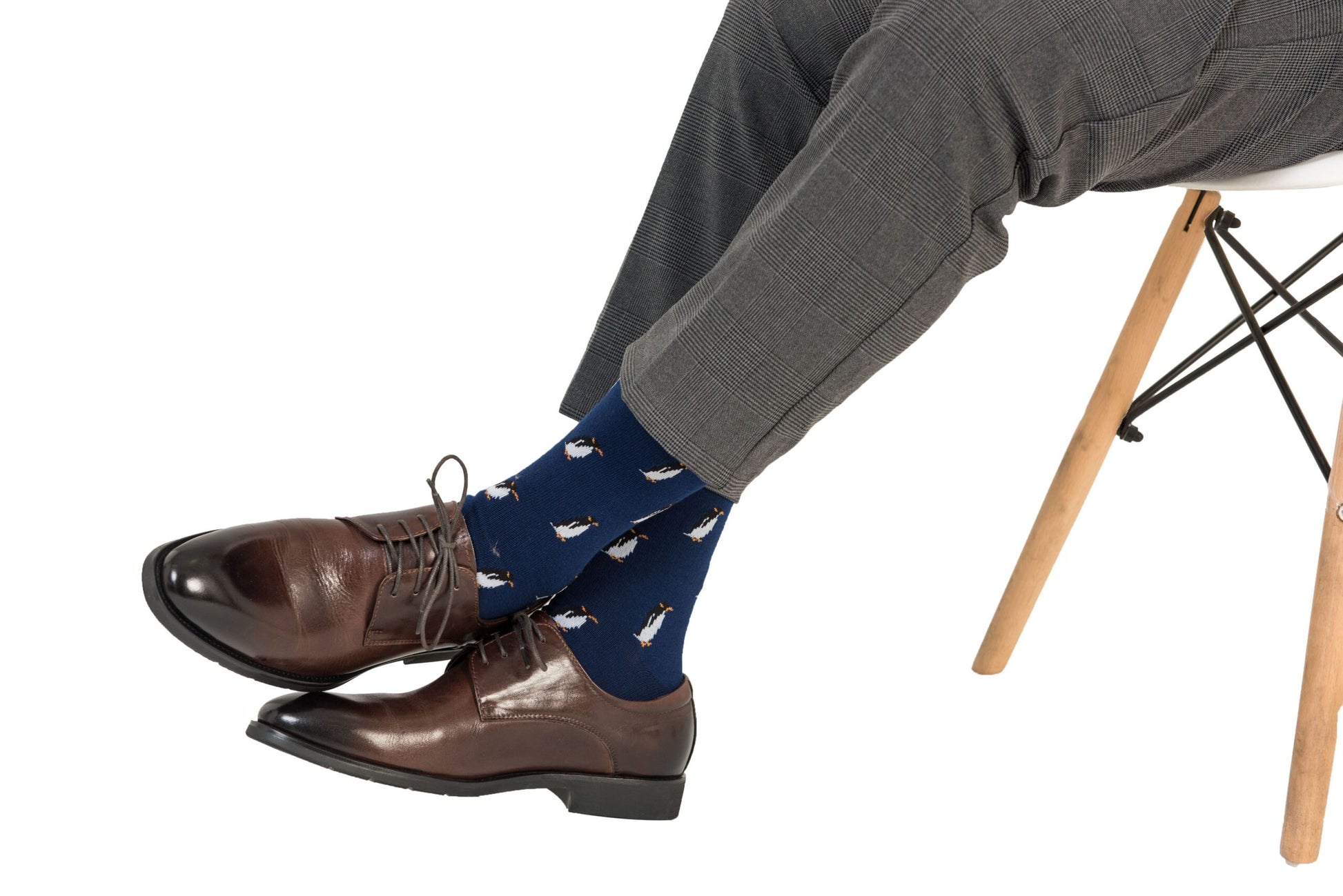 Person wearing brown leather dress shoes and Penguin Socks, seated on a white chair with light wooden legs, exuding Arctic allure.