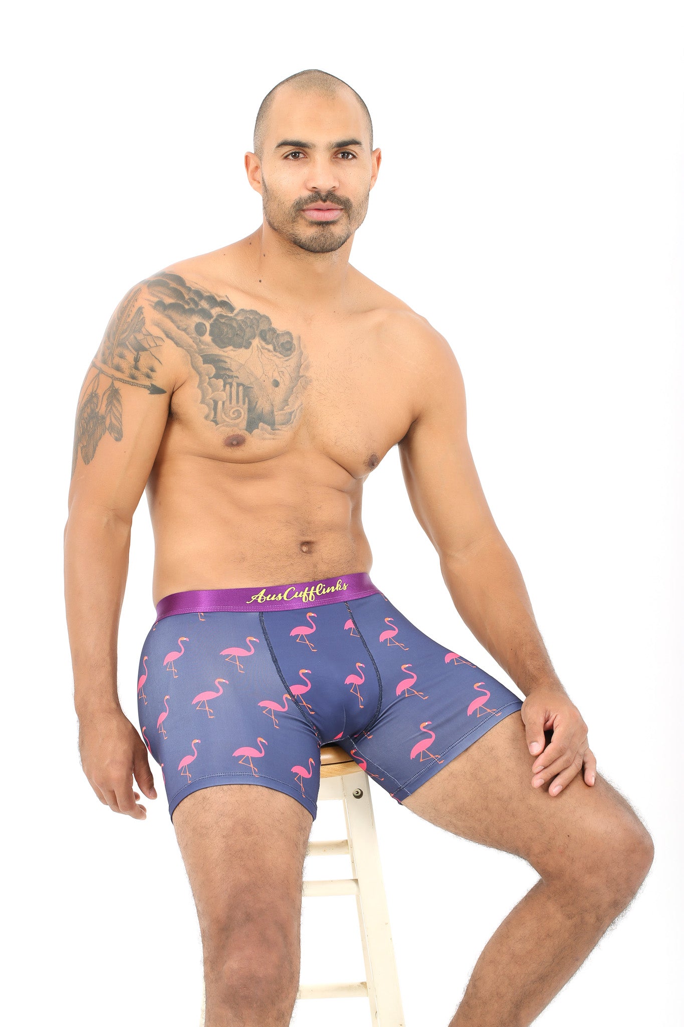 A bald man with a chest tattoo, wearing Blue Boxer Briefs with the Pink Flamingo Underwear pattern, sits on a white stool against a white background.