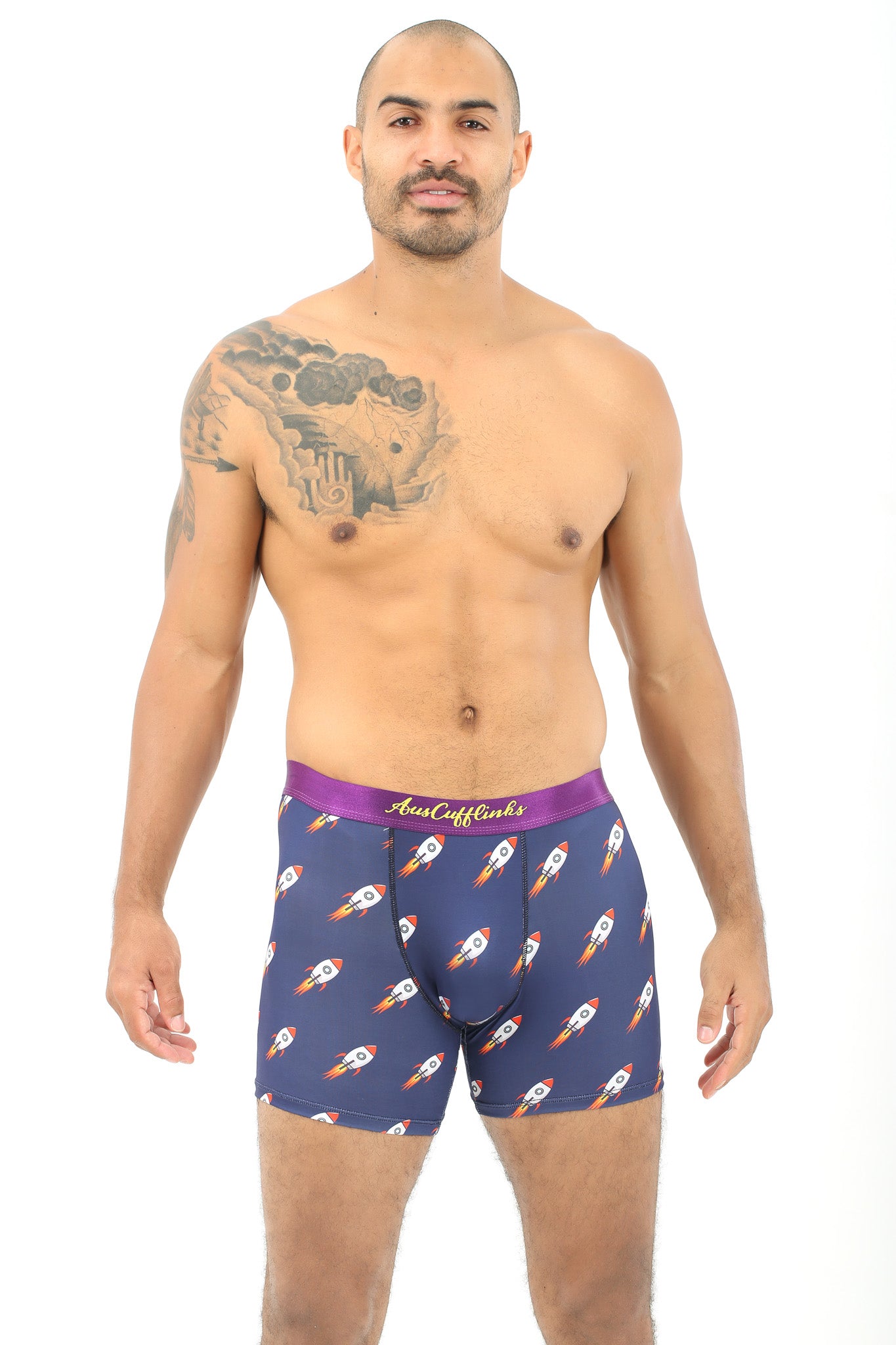 A man with a tattoo on his left arm is modeling Rocket Underwear, featuring blue boxer briefs adorned with rocket ship patterns for a delightful blend of comfort and cosmos-themed style.