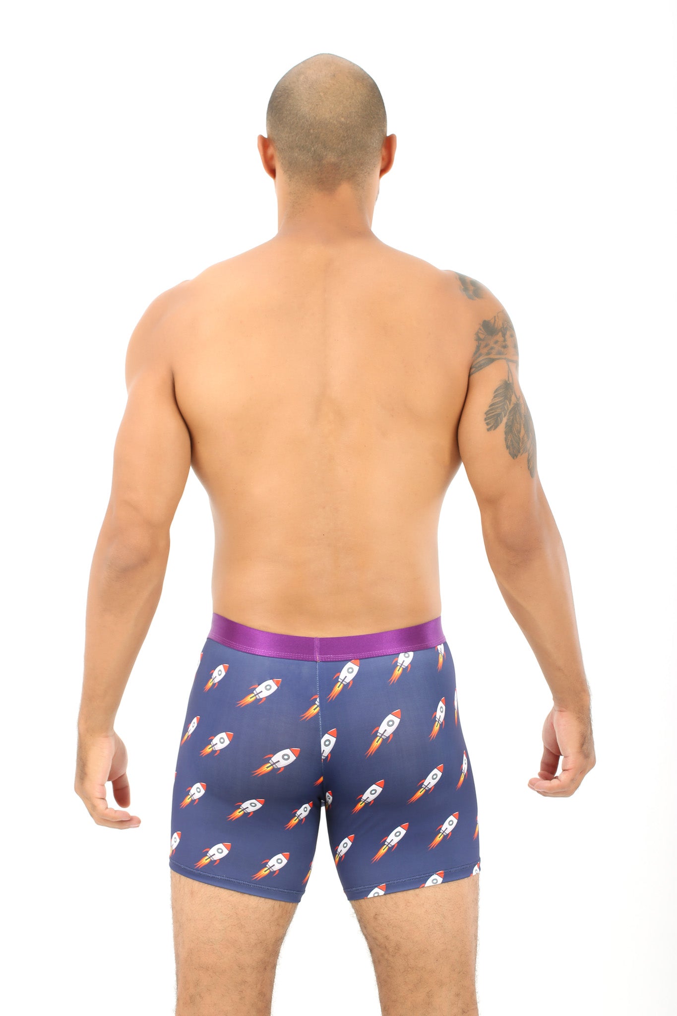 A person with a shaved head and a tattoo on their left arm stands facing away, embracing the comfort of their cosmic-themed Rocket Underwear, adorned with rocket ship designs.