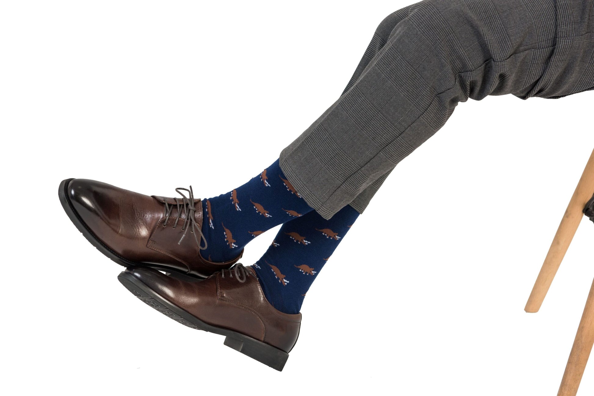 A person with panache wears brown leather shoes, Triceratops Socks in blue featuring orange dog patterns, and gray checkered pants, as they sit with their legs crossed on a chair.