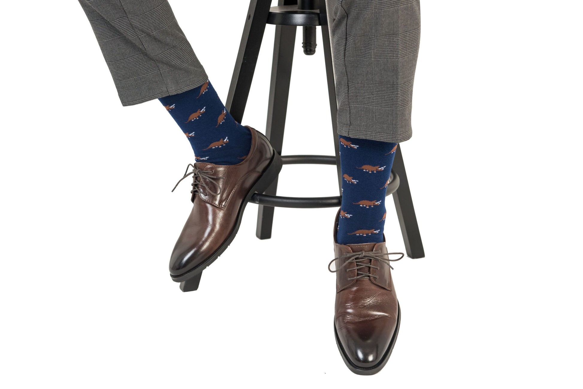 A person sitting on a stool wearing gray pants, Triceratops Socks featuring blue hues with animal patterns, and brown shoes exuding a touch of panache.