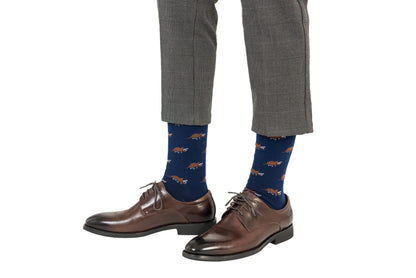 A person exudes prehistoric panache, sporting gray plaid pants, Triceratops Socks in blue adorned with dinosaur patterns, and brown leather shoes.