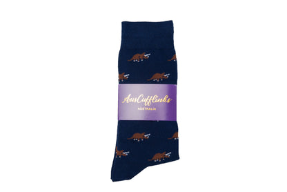 Triceratops Socks feature a navy blue design embellished with a brown triceratops dinosaur pattern, offering a touch of prehistoric charm, and come wrapped in a purple label with cursive text.