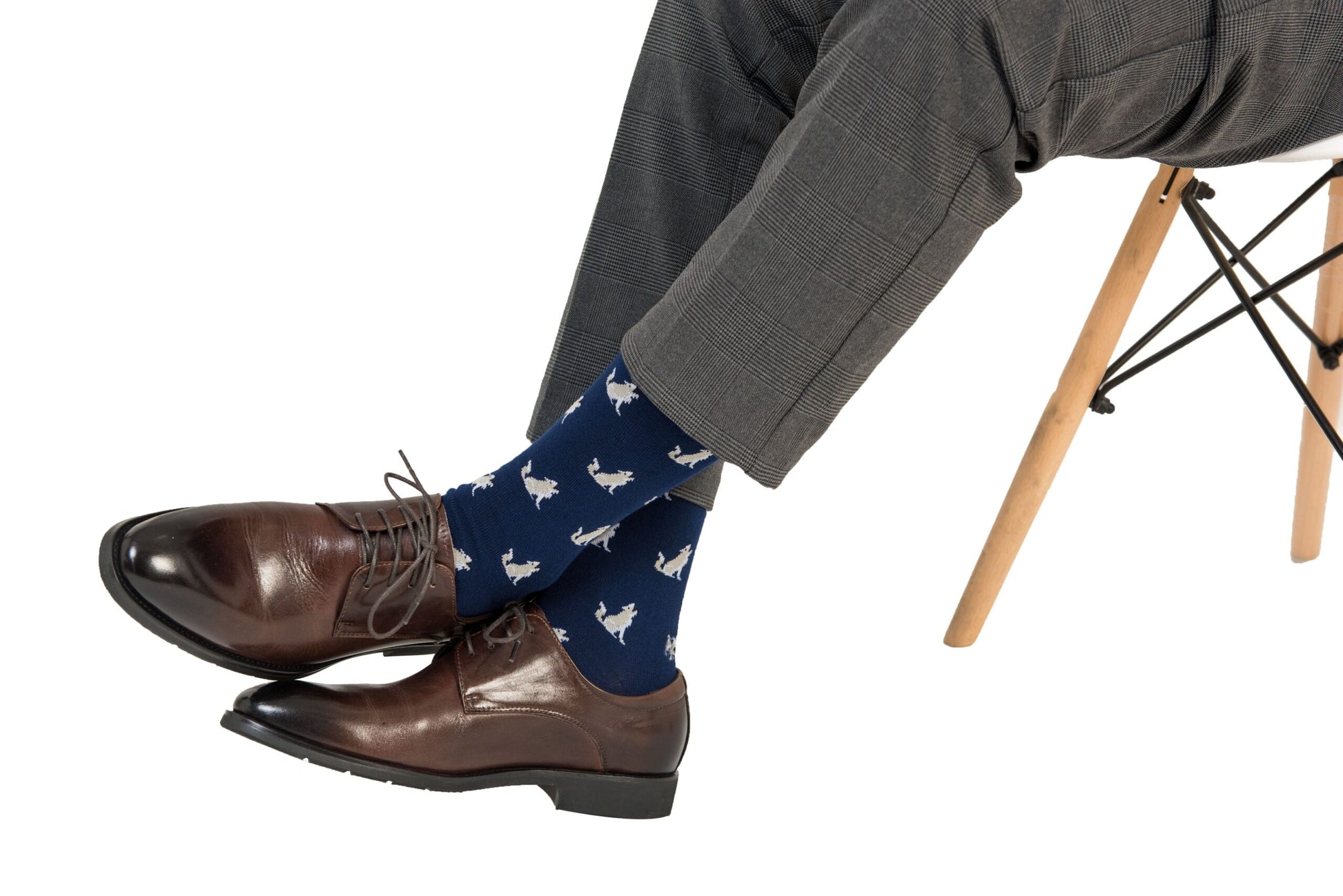 Man in grey trousers and brown shoes, leading the pack with every step, sports Wolf Socks adorned with a white bird pattern, seated with one leg crossed over the other.