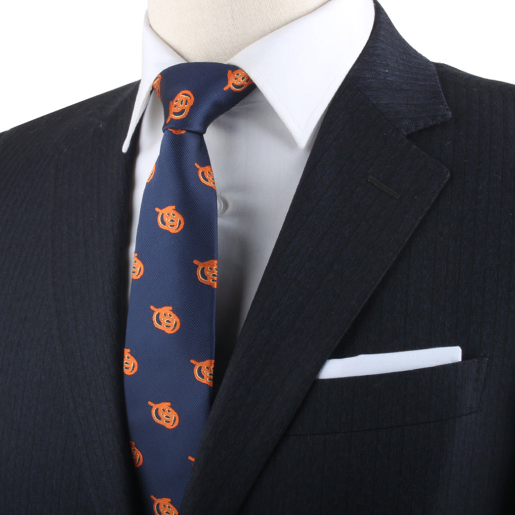 A man in a formal black suit with a white shirt and the Pretzel Skinny Tie, adorned with orange pumpkin designs, exudes charisma. A white pocket square is neatly placed in the jacket's pocket, adding a charming twist to his ensemble.