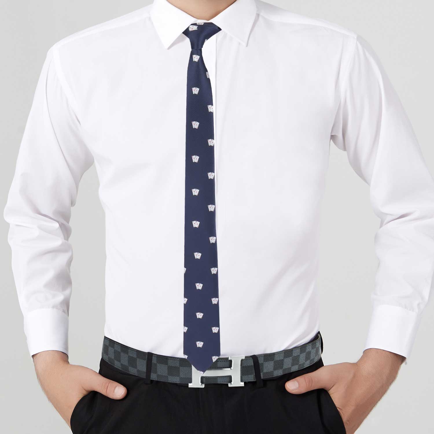 A person wearing a white dress shirt, the Ace of Spades Tie featuring a playful card pattern, and black trousers with a checkered belt and silver buckle stands with their hands in their pockets. This unique touch truly elevates your style.