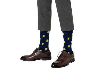A person wears gray plaid trousers, brown leather dress shoes, and navy blue Beer Socks with yellow beer mug patterns—Beer Socks are fun socks perfect for beer lovers—shown on a white background.