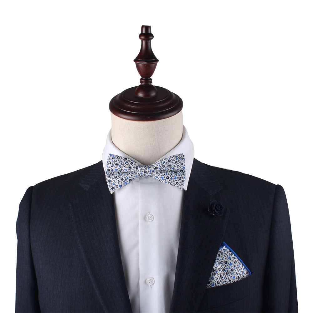 A mannequin dressed in a dark suit with a white shirt, adorned with the Black Light Blue Floral Cotton Bow Tie and Pocket Square Set, and a coordinating floral lapel pin.