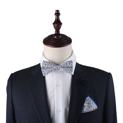 A mannequin dressed in a dark suit with a white shirt, adorned with the Black Light Blue Floral Cotton Bow Tie and Pocket Square Set, and a coordinating floral lapel pin.