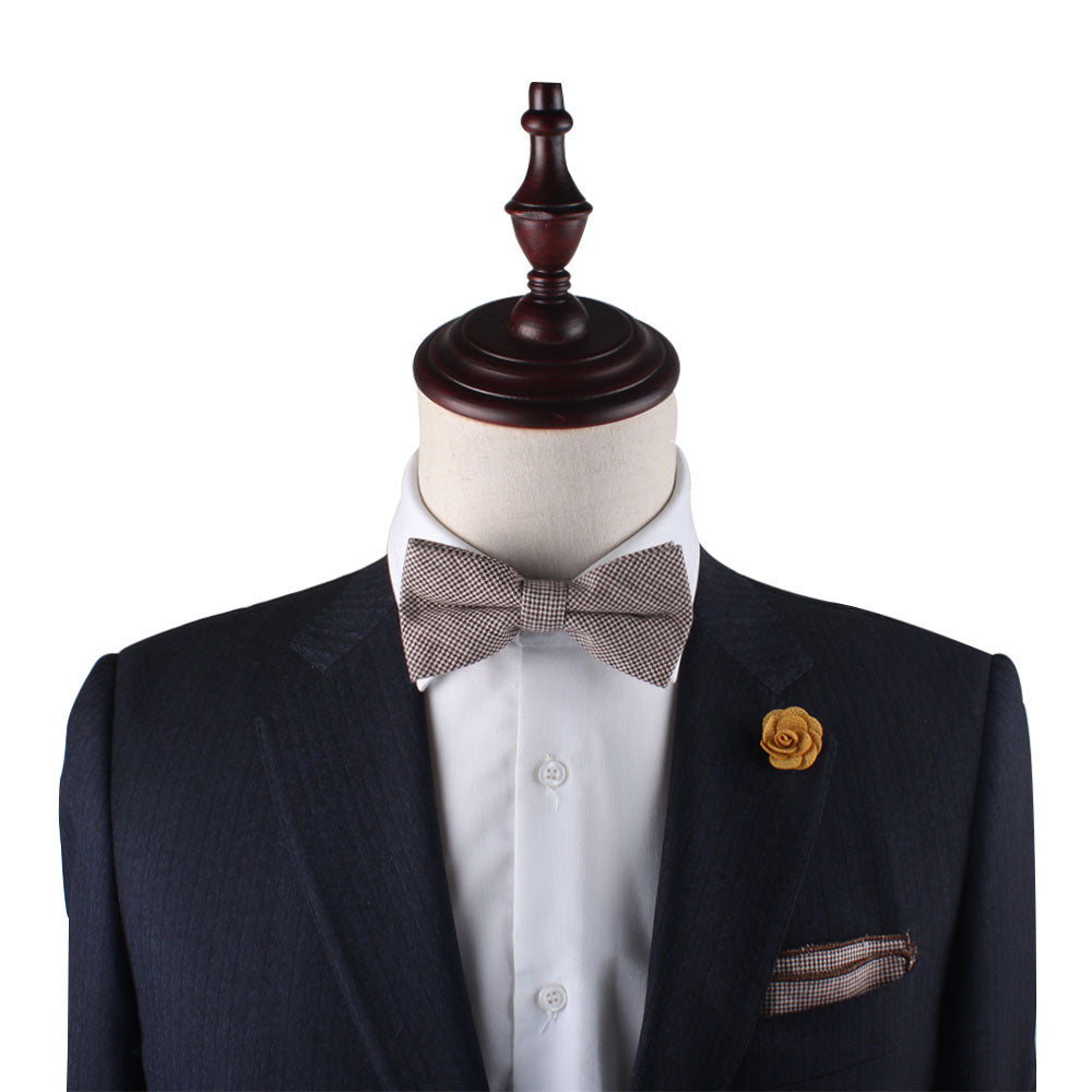 A mannequin sports a dark suit jacket with a white shirt, a checkered bow tie, and a yellow flower lapel pin exuding timeless elegance. The sophisticated ensemble is enhanced by the classic touch of the Brown Mini Houndstooth Cotton Bow Tie & Pocket Square Set for refined style lovers.